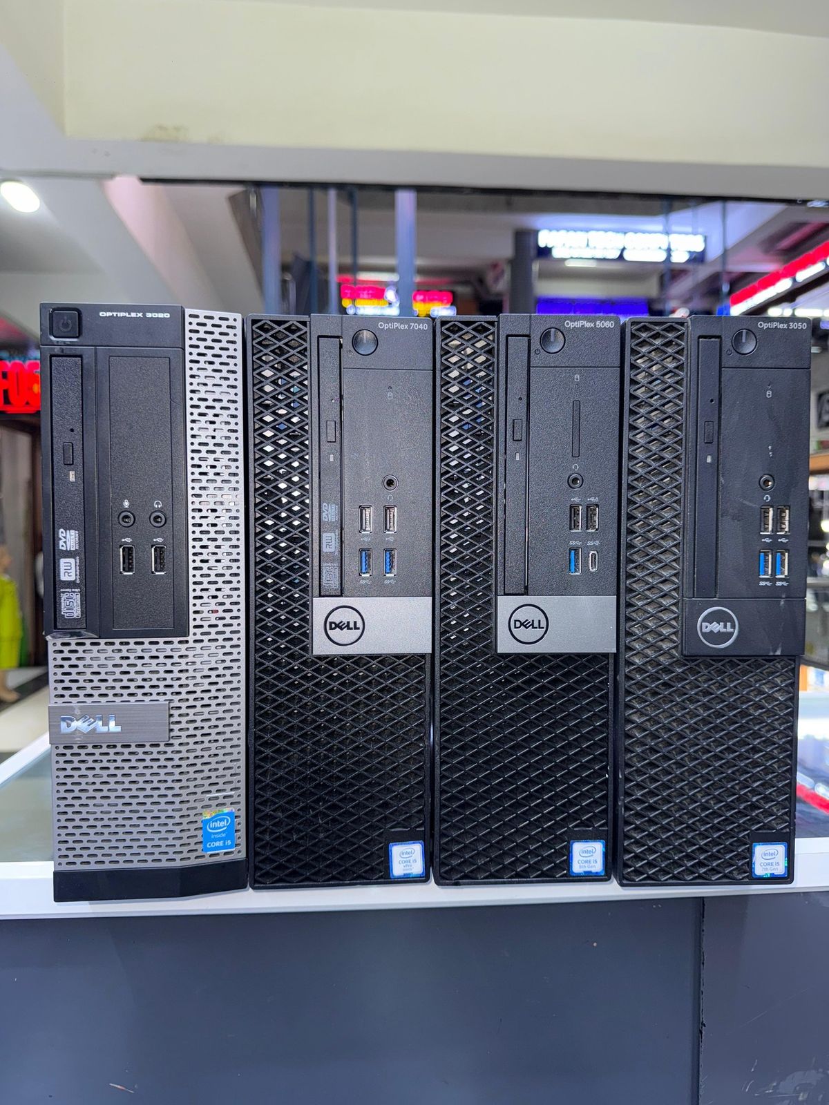 Four Dell desktop towers in different models and colors (silver, black, and dark gray) lined up on a shelf in what appears to be a retail or office space.